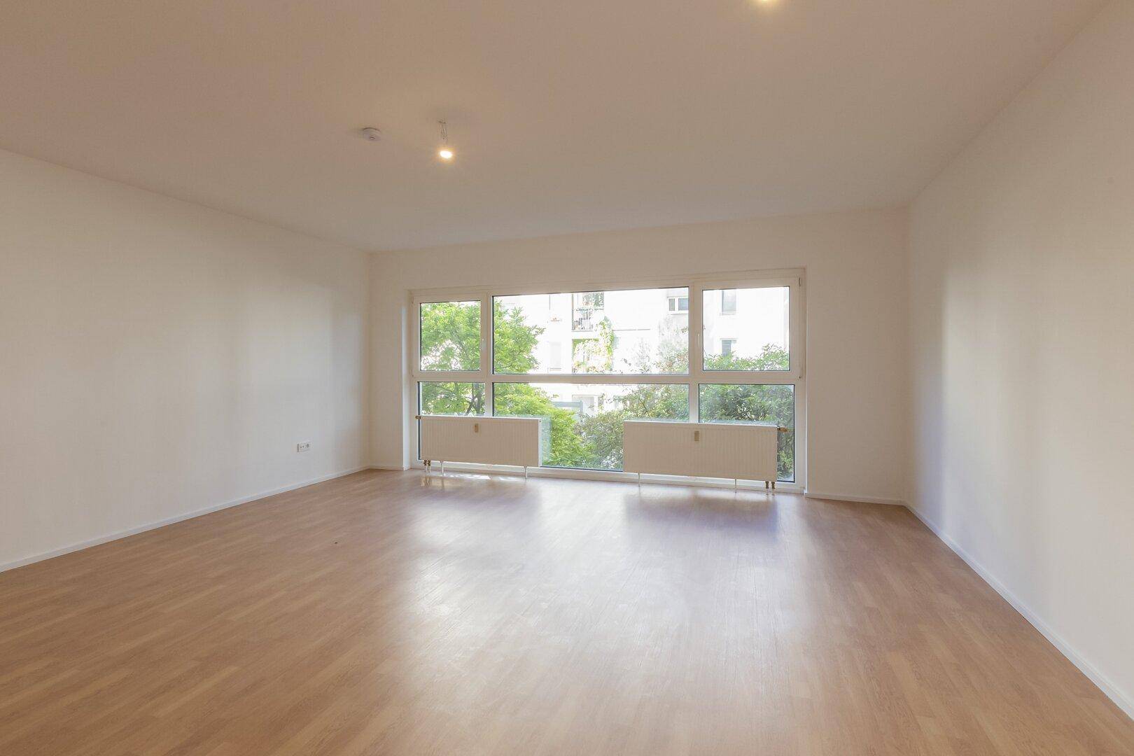 Geräumiges Wohnzimmer mit viel Tageslicht, hellem Laminatboden und neutralen Wänden.