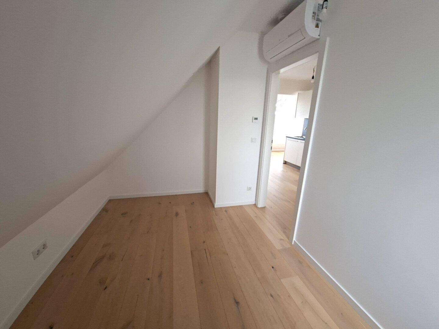 Heller Raum mit Holzboden und Zugang zu einem weiteren Zimmer, Klimaanlage sichtbar.