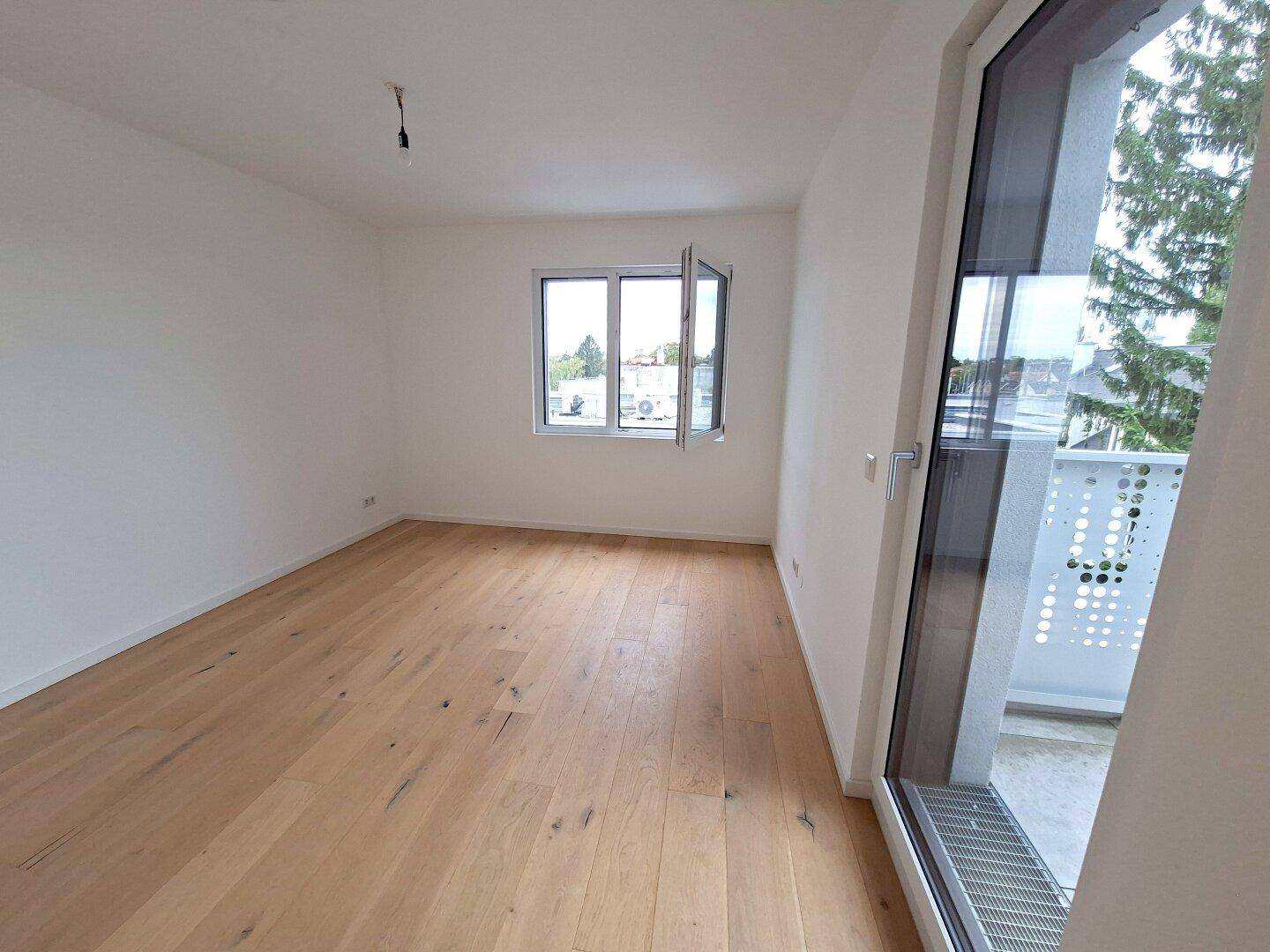Geräumiger, heller Raum mit Holzboden und Zugang zu einem Balkon oder einer Terrasse.