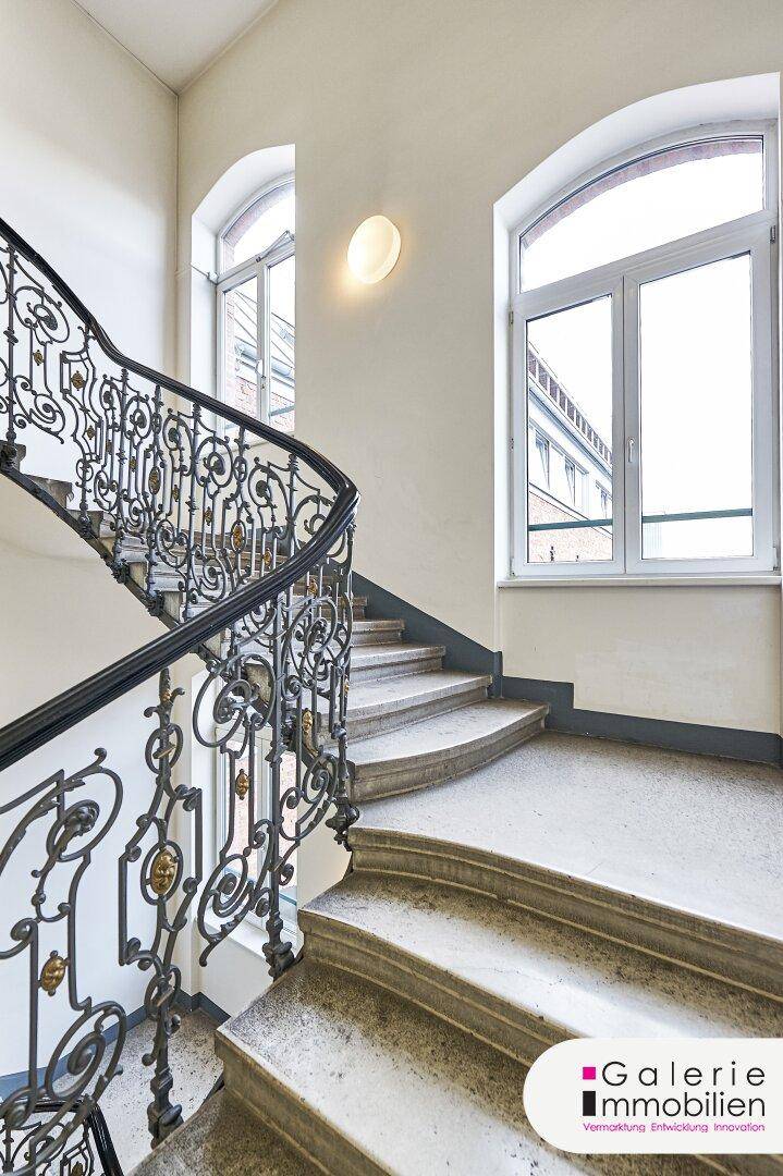 Helles Treppenhaus mit elegantem schmiedeeisernem Geländer und großen Rundbogenfenstern, typisch für Altbauten.