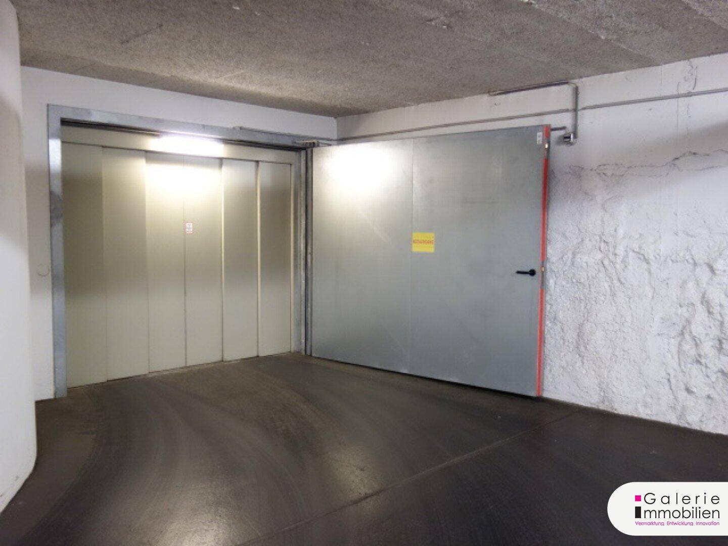 Zugang zu einem Aufzug und einer Metalltür innerhalb der Tiefgarage, mit rauen Wänden und Beleuchtung.