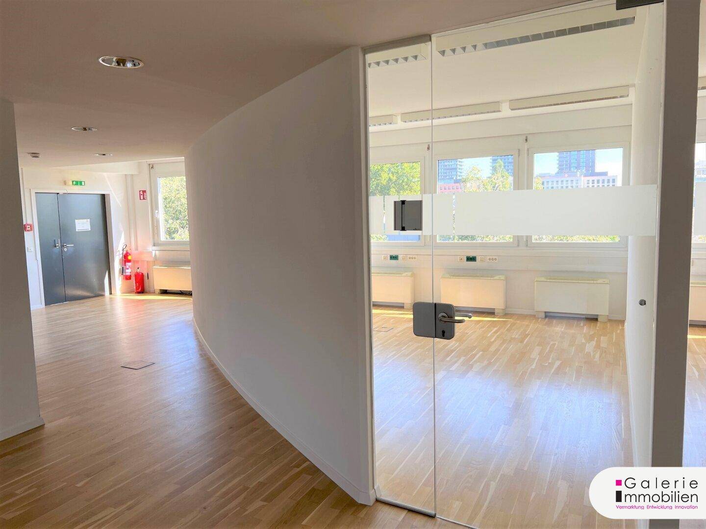 Langer, heller Flur mit Holzboden und einer modernen Glastür, die zu einem Büro führt und Offenheit vermittelt.