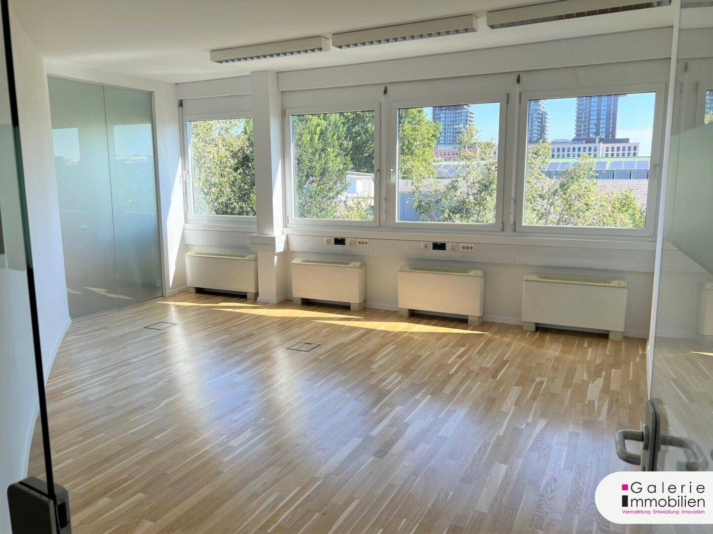 Modernes Büro mit Glaswänden, Holzboden und großen Fenstern, die viel Licht spenden und eine offene Atmosphäre schaffen.