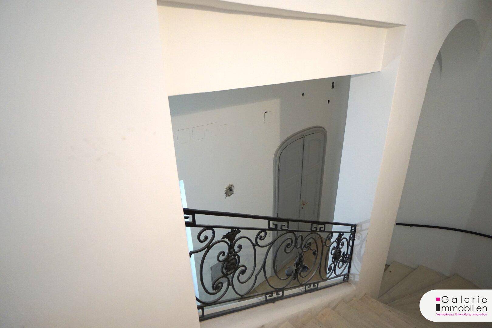 Blick in ein Treppenhaus mit kunstvoll geschmiedetem Geländer und weiß gestrichenen Wänden.