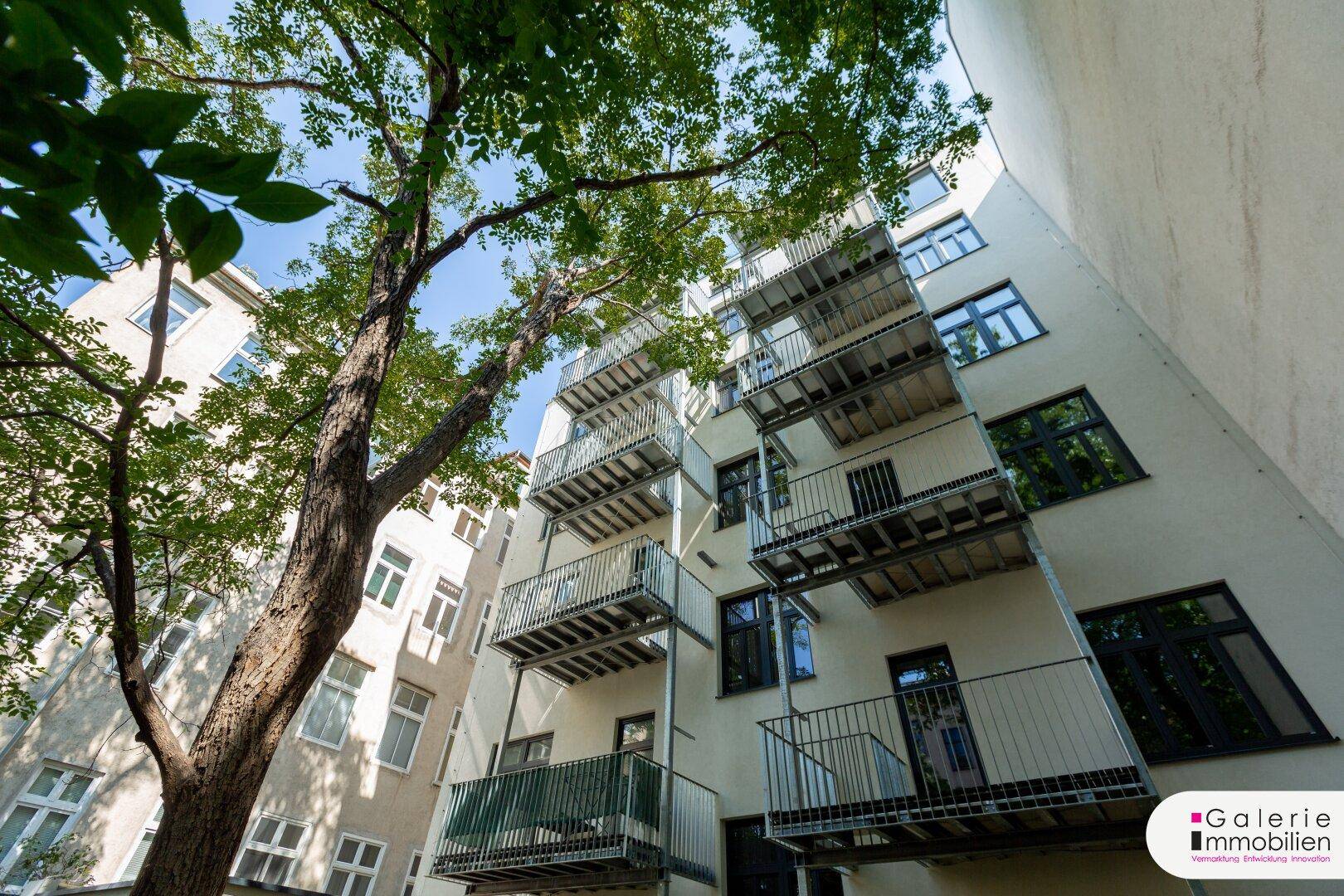 Moderne Gebäudefassade mit gestapelten Balkonen und einem großen Baum im Vordergrund.