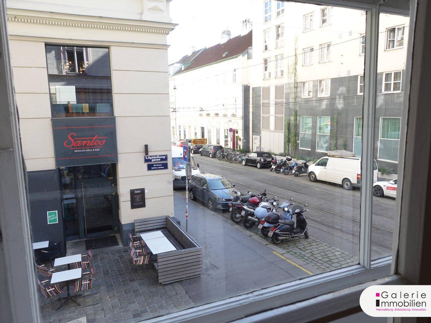 Blick aus dem Fenster auf eine belebte Straße mit Geschäften und parkenden Fahrzeugen.