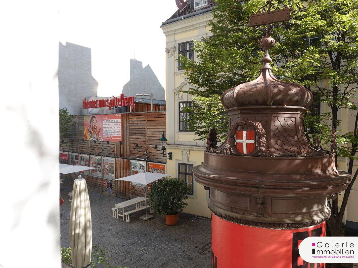Blick auf einen belebten Außenbereich mit Bäumen, Sitzgelegenheiten und einer historischen Säule.
