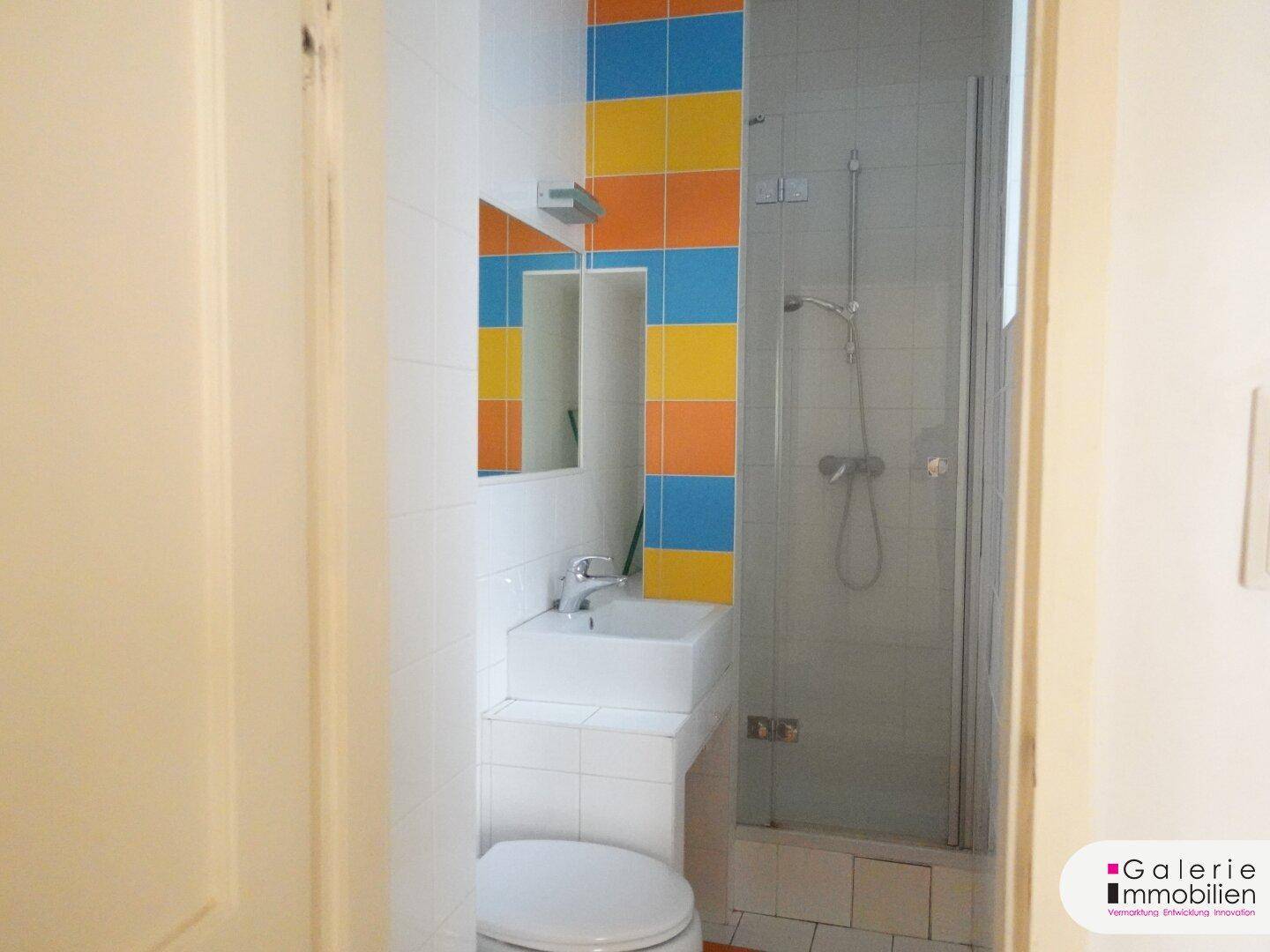 Kompaktes Badezimmer mit Duschkabine, WC und Waschbecken, akzentuiert durch bunte Fliesen.