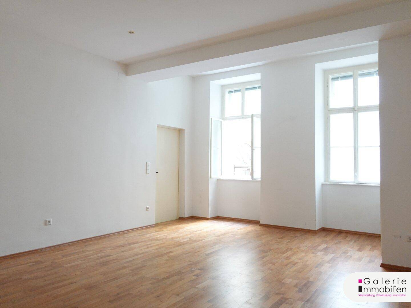 Heller, leerer Raum mit Parkettboden und zwei großen Fenstern, die viel Tageslicht hereinlassen.