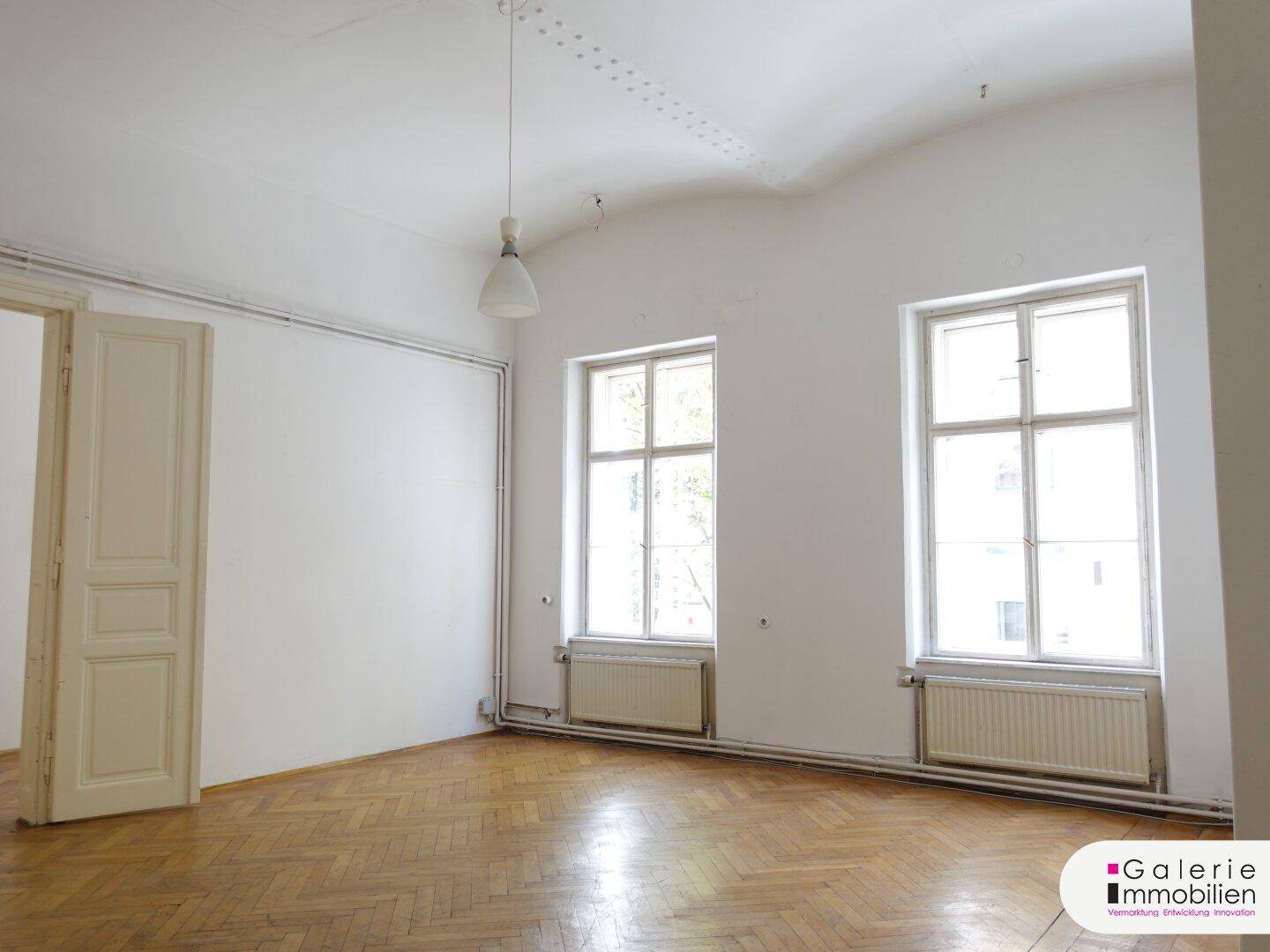 Geräumiger, heller Raum mit Fischgrätparkett, hohen Fenstern und Heizkörpern unter den Fenstern.