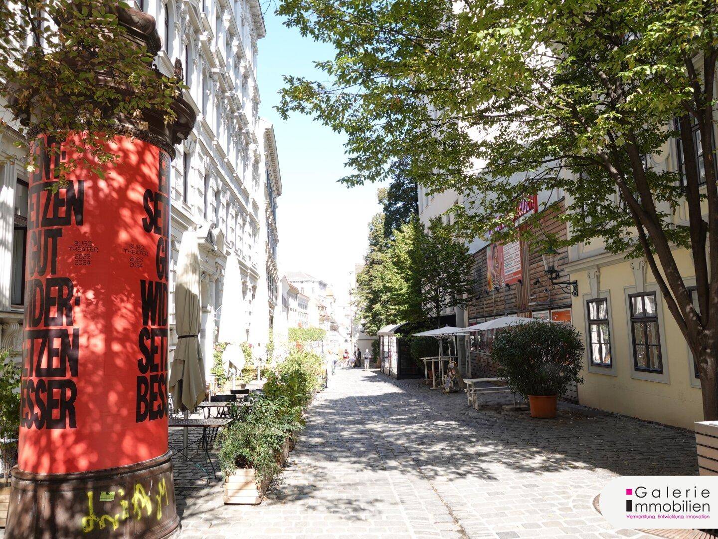 Historische Gasse mit Kopfsteinpflaster, Bäumen und einer markanten roten Säule im Vordergrund.