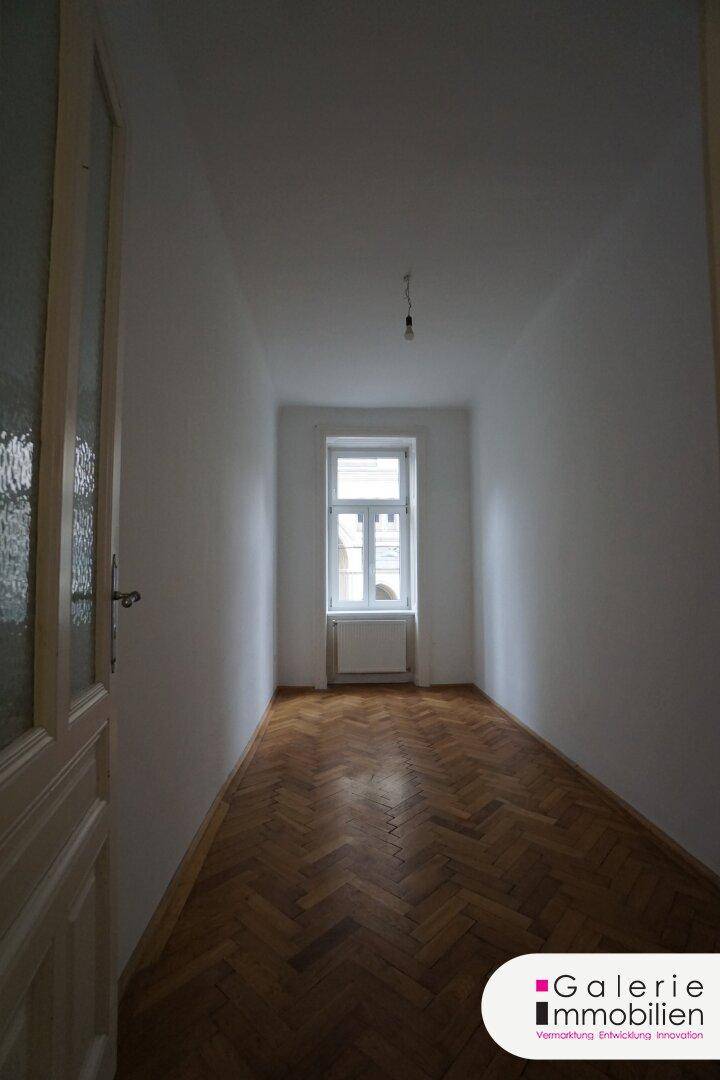 Schmaler Raum mit Fischgrätparkettboden und einem Fenster, das Tageslicht hereinlässt.