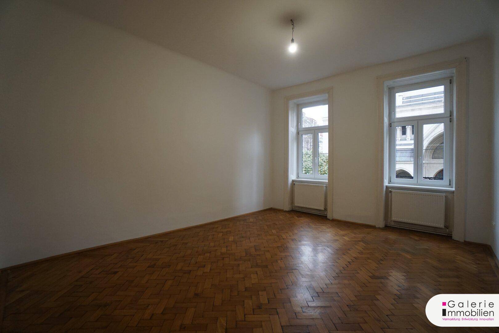 Heller Raum mit Fischgrätparkettboden und zwei großen Fenstern mit Blick auf den Innenhof.