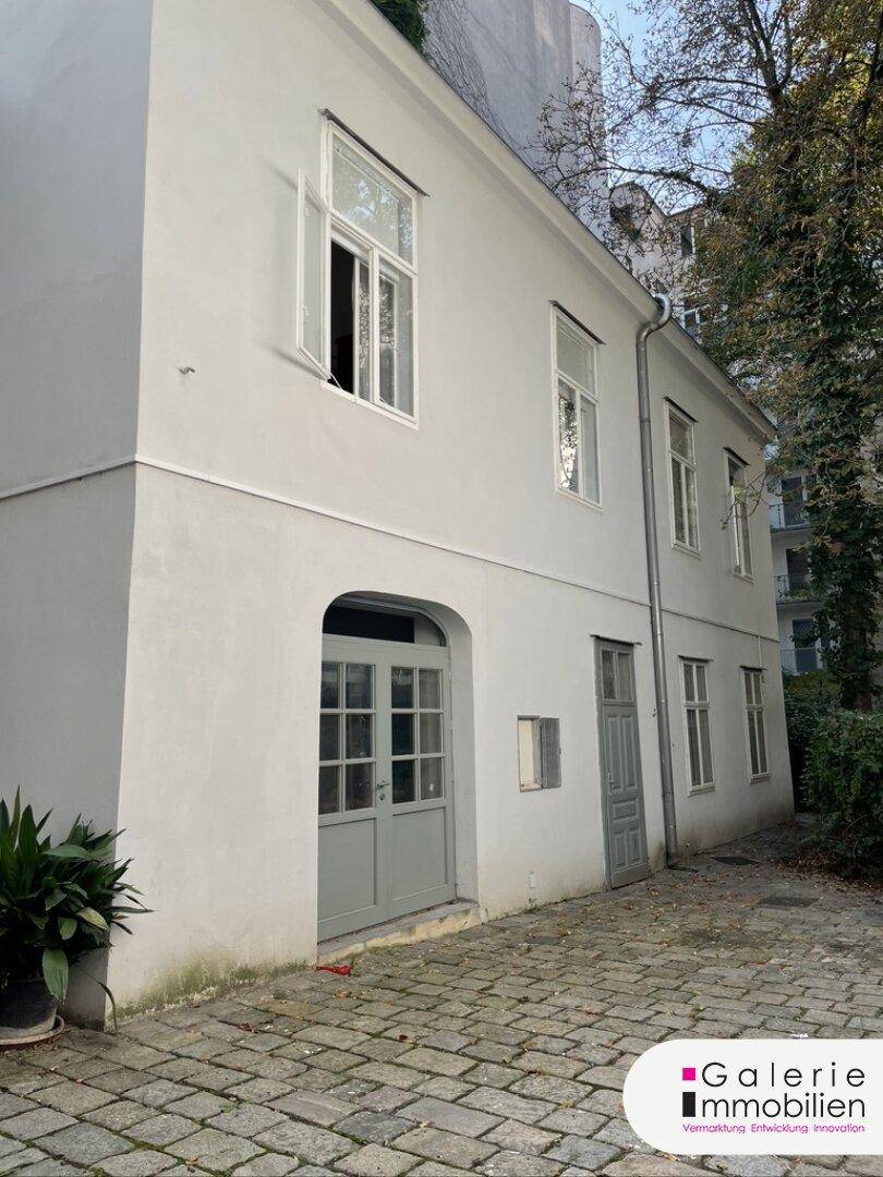 Klassisches weißes Gebäude mit gepflastertem Innenhof und hohen Fenstern, umgeben von Bäumen.