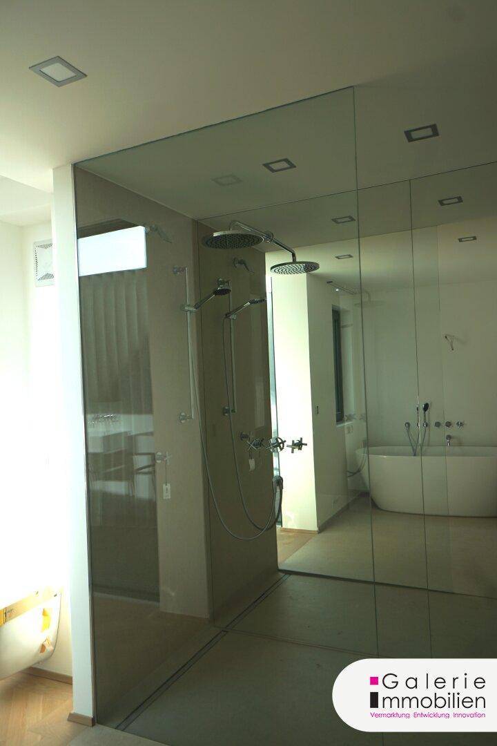 Modernes Badezimmer mit großer Spiegelfläche, Regendusche und freistehender Badewanne im Hintergrund.