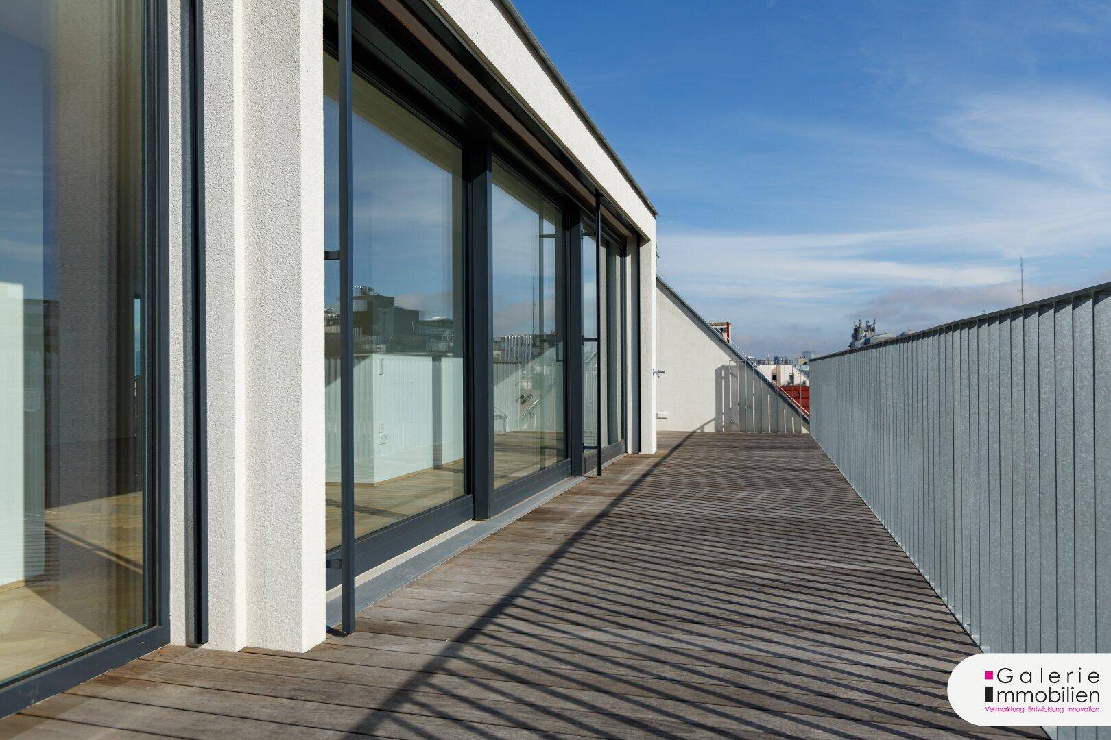 Lange Terrasse mit Holzboden und Metallgeländer, die sich entlang der modernen Fassade erstreckt.