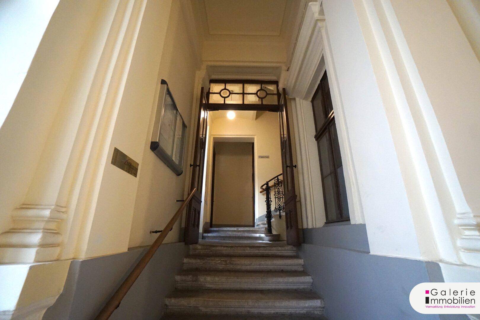 Historischer Eingangsbereich mit Steintreppe, schmiedeeisernem Geländer und hohen Decken.