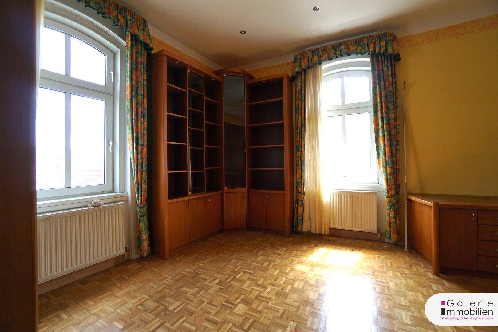 Raum mit Parkettboden, zwei Fenstern und einem großen eingebauten Bücherregal.