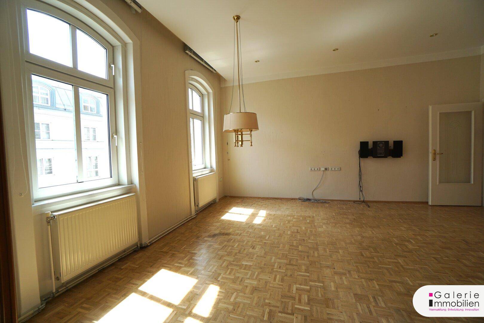 Helles Wohnzimmer mit Parkettboden, großen Fenstern und einer Wandhalterung für Elektronik.