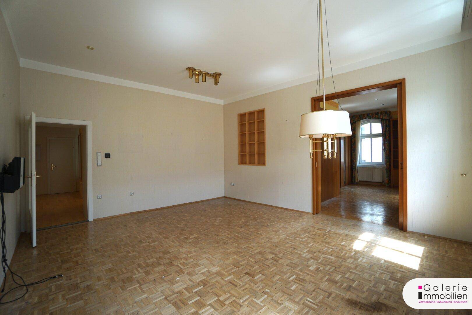 Geräumiges Wohnzimmer mit Parkettboden, zwei Fenstern und mehreren Türöffnungen.