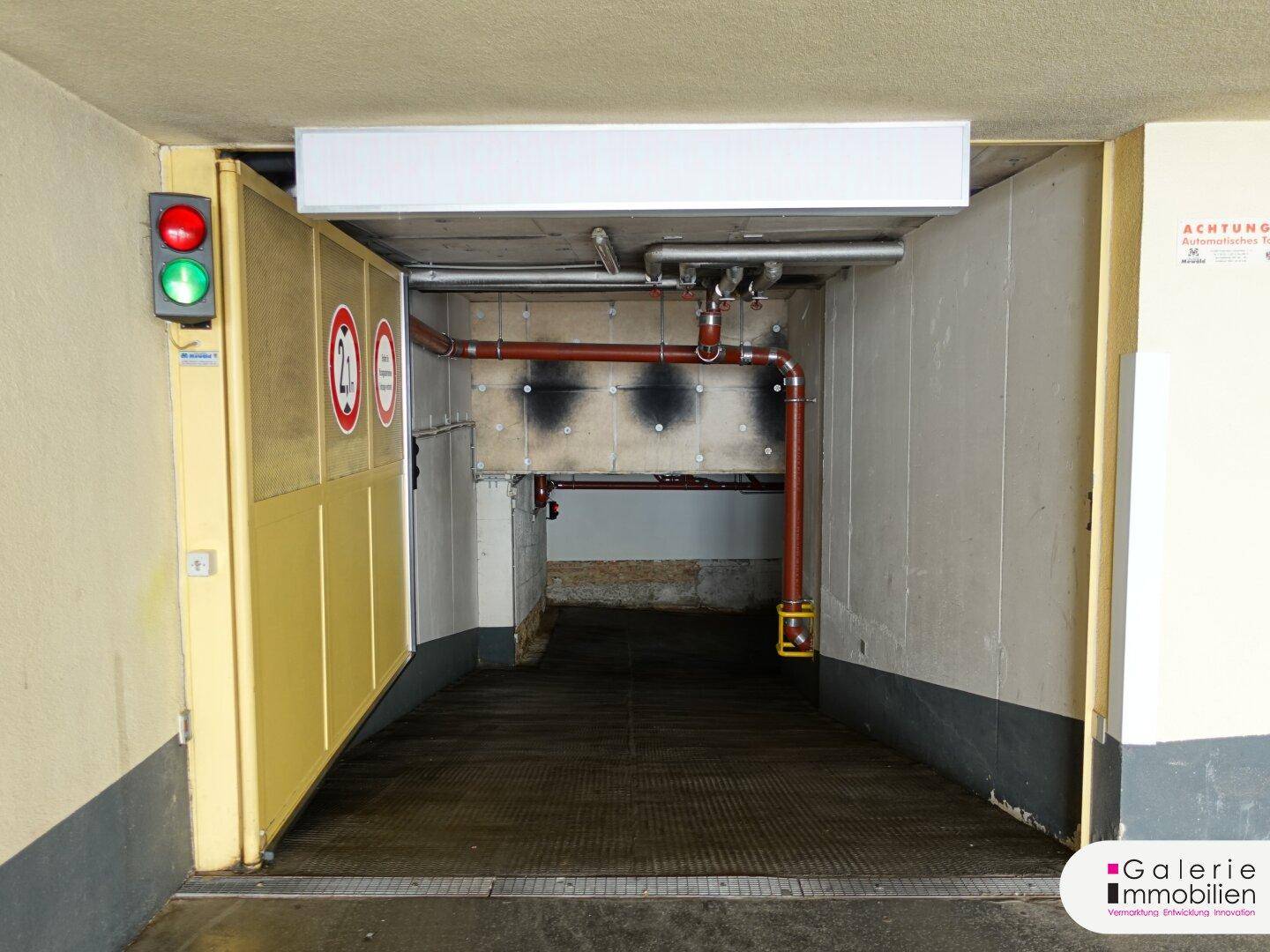 Blick in den Einfahrtsbereich der Tiefgarage mit geöffnetem Tor, Rampe und sichtbaren Leitungen.