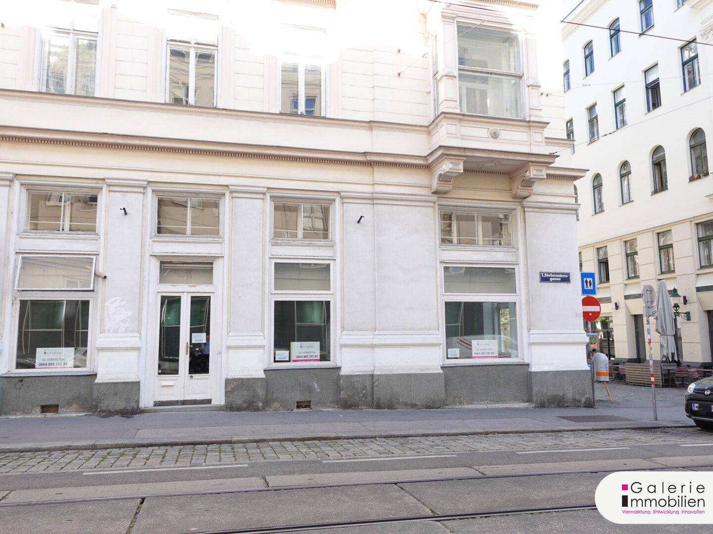Die helle Fassade des Gebäudes mit großen Schaufenstern und einem Erker im oberen Stockwerk.
