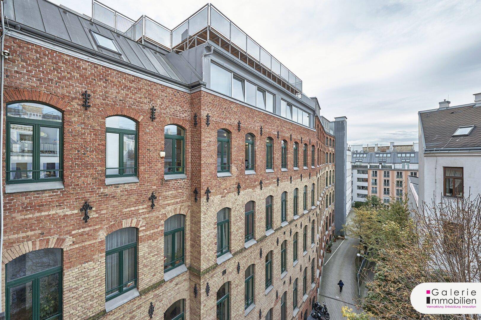 Historische Backsteinfassade eines Gebäudes mit grünen Fensterrahmen und moderner Dachterrasse.