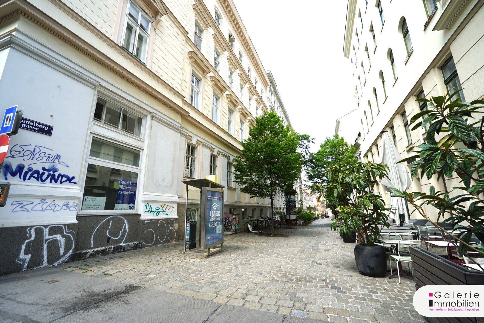 Kopfsteingepflasterte Gasse mit Bäumen, Sitzgelegenheiten und Gebäuden im urbanen Umfeld.