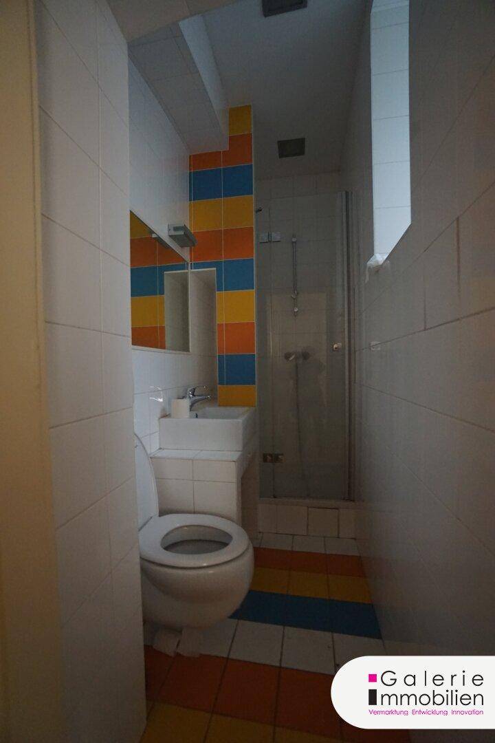 Kleines Badezimmer mit Dusche, WC und farbigen Fliesen in Blau, Orange und Gelb.
