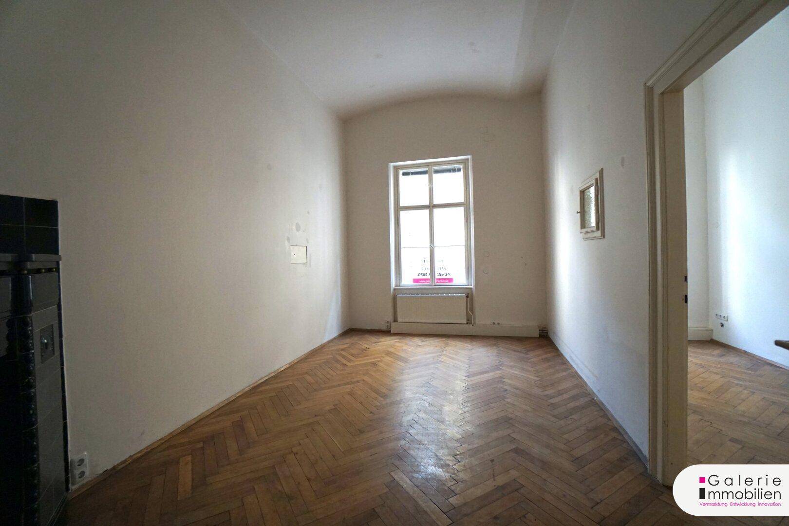 Geräumiges Zimmer mit Fischgrätparkett, Fenster und Blick auf den Kachelofen.