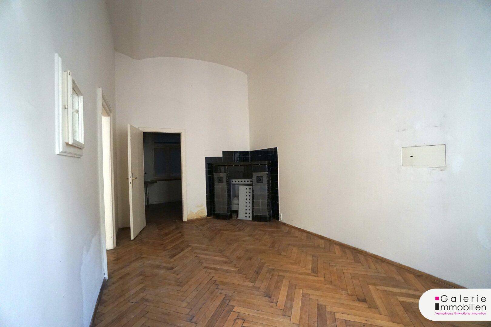 Helles Zimmer mit Fischgrätparkett und einem traditionellen Kachelofen an der Wand.