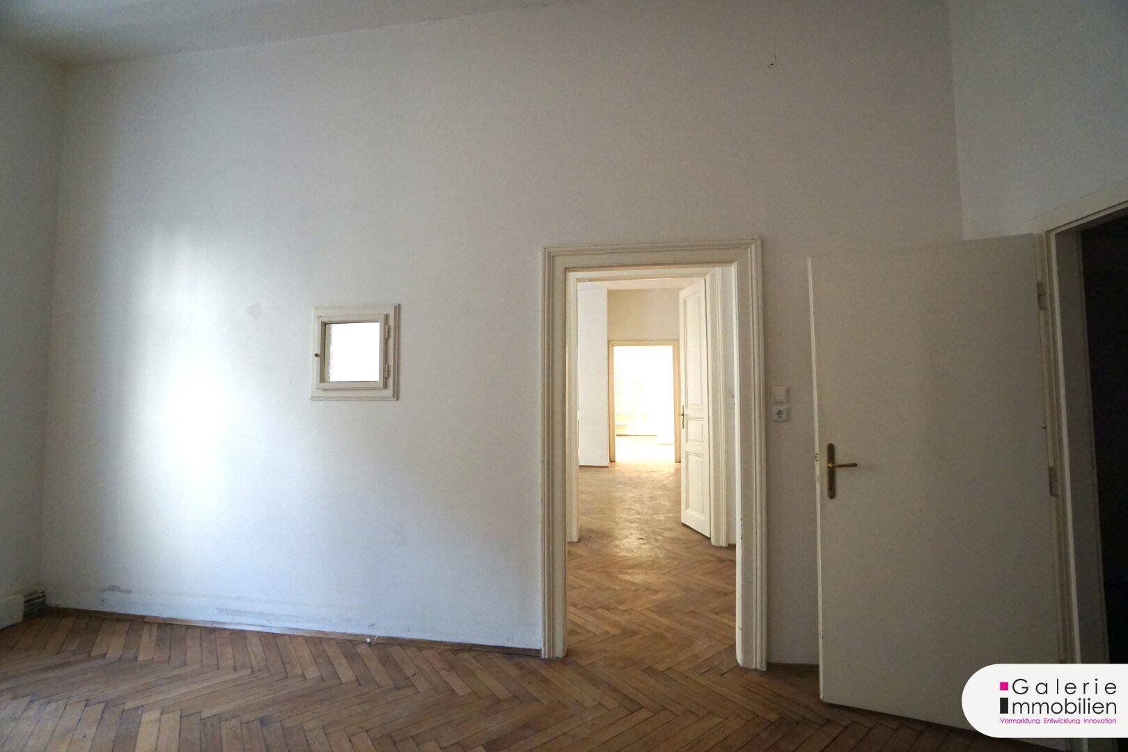 Helles Zimmer mit Fischgrätparkett und Zugang zu weiteren Räumen durch offene Türen.