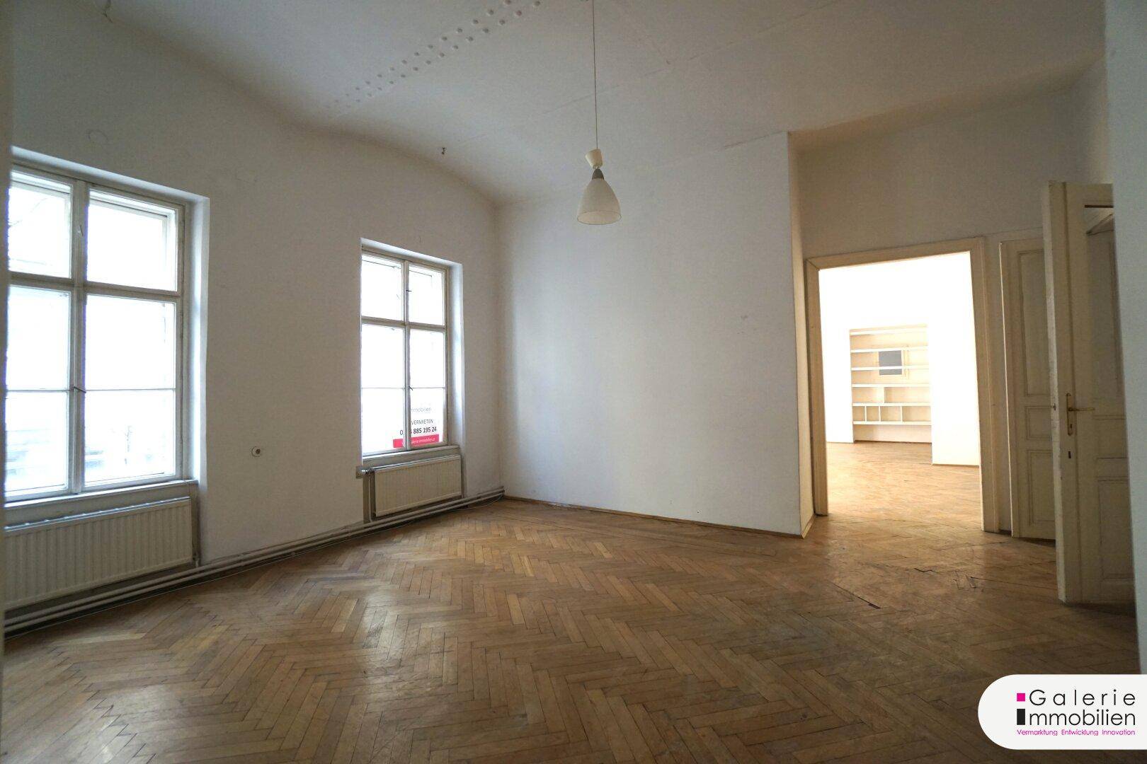 Helles Zimmer mit Fischgrätparkett, hohen Decken und Blick auf den angrenzenden Raum.