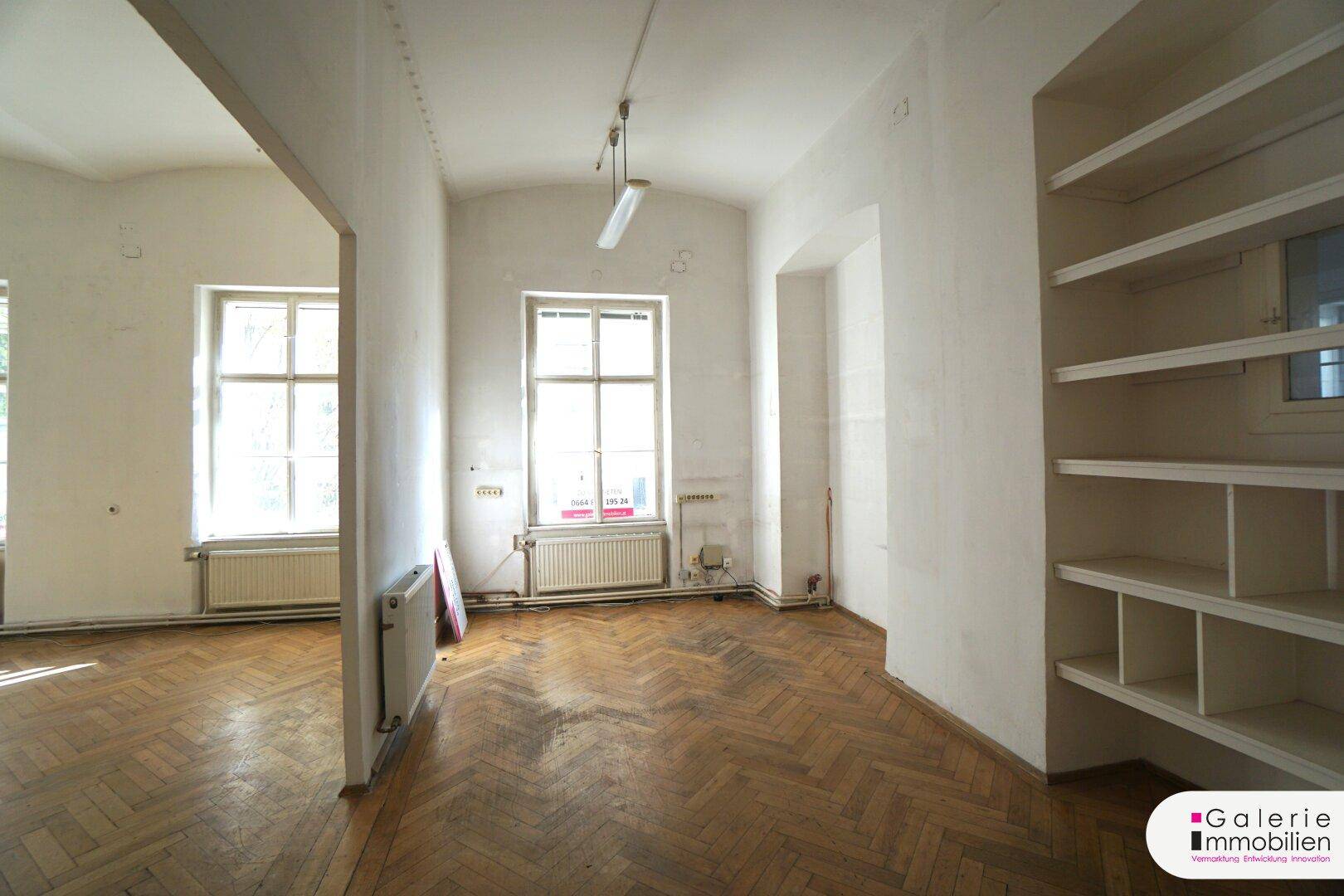 Geräumiges Zimmer mit Fischgrätparkett, hohen Decken und zwei großen Fenstern, bereit zur Renovierung.