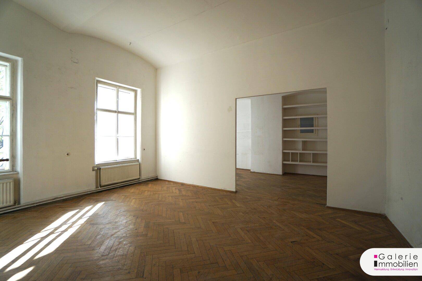 Geräumiges Zimmer mit Fischgrätparkett und viel Tageslicht, das auf den Boden fällt.