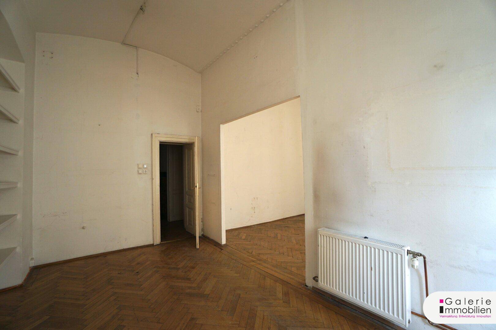 Geräumiges Zimmer mit Fischgrätparkett, hohen Decken und Zugang zu einem weiteren Raum.