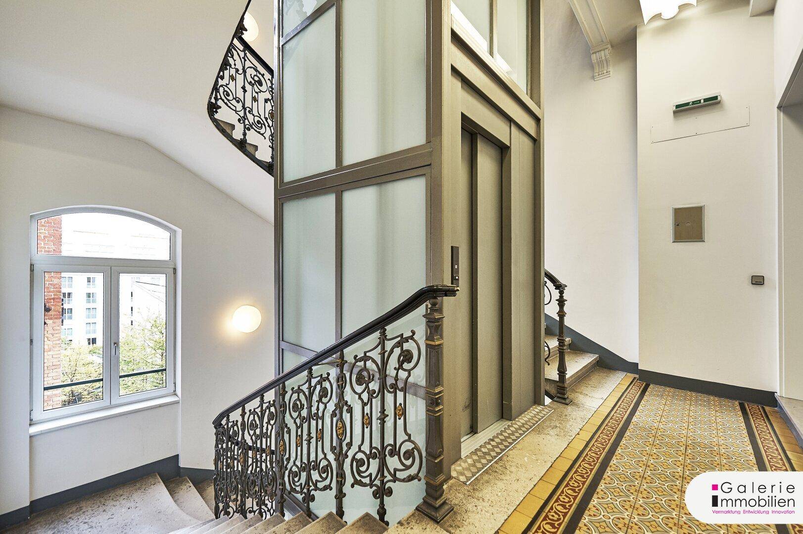 Historisches Treppenhaus mit schmiedeeisernem Geländer, modernem Aufzug und hohen Decken.