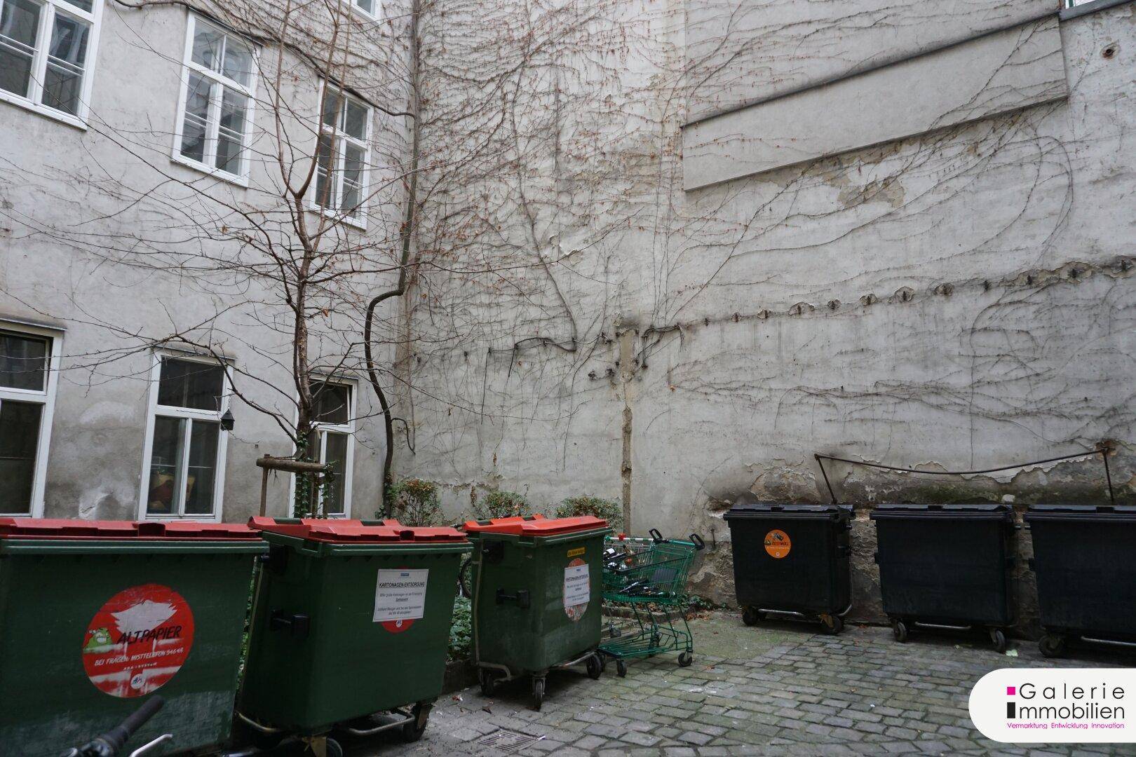 Hinterhof mit alten Gebäudewänden, spärlicher Vegetation und Mülltonnen.
