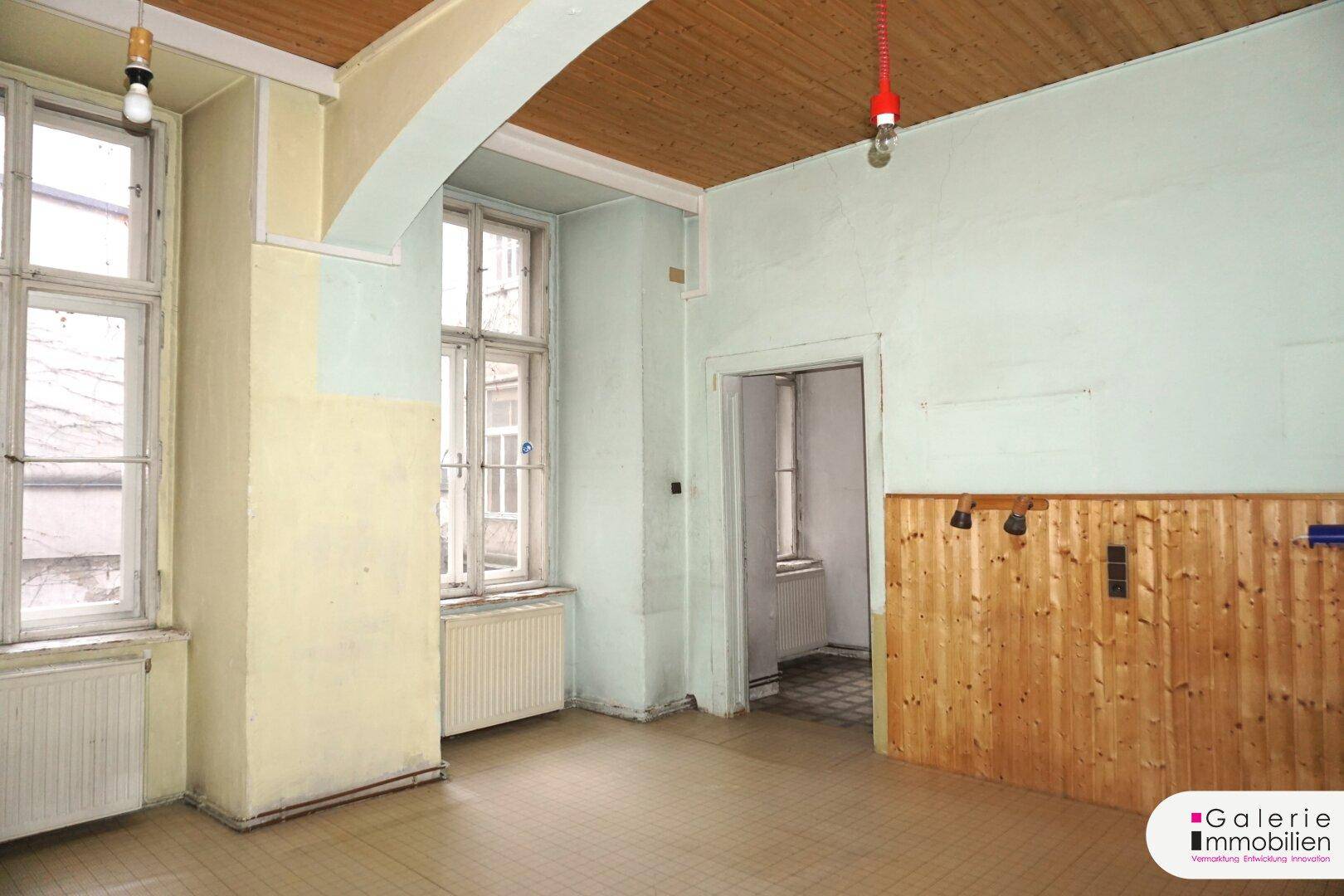 Renovierungsbedürftiger Raum mit Holzbalkendecke, zwei Fenstern und Holzvertäfelung an einer Wand.
