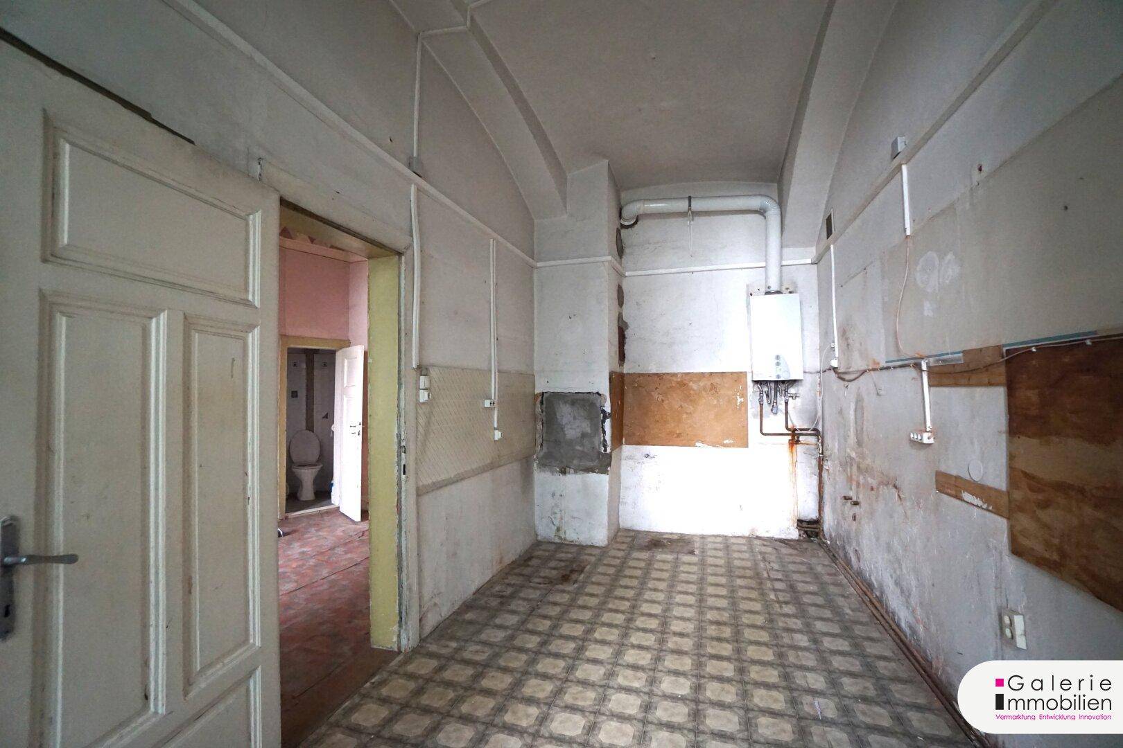 Renovierungsbedürftiger Raum mit sichtbaren Rohren und einem Durchgang zu einem WC, ideal für eine Küche.