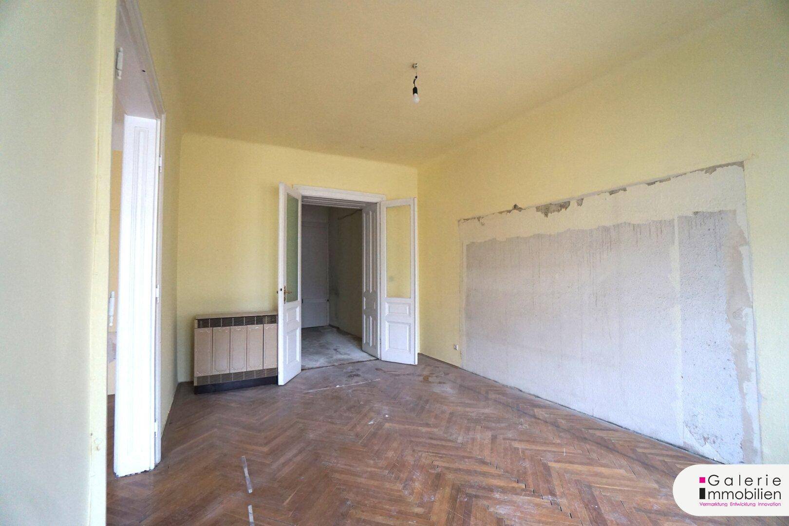 Geräumiges Zimmer mit Fischgrätparkett und sichtbaren Spuren früherer Wandgestaltung.