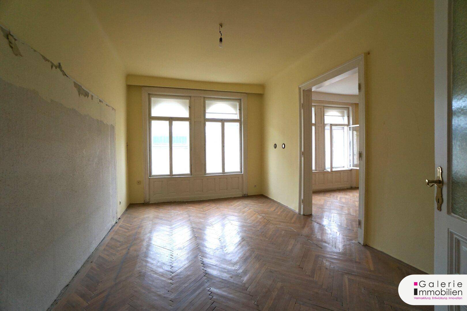 Großes Zimmer mit Fischgrätparkett, hohen Fenstern und Durchgang zu einem weiteren Raum.