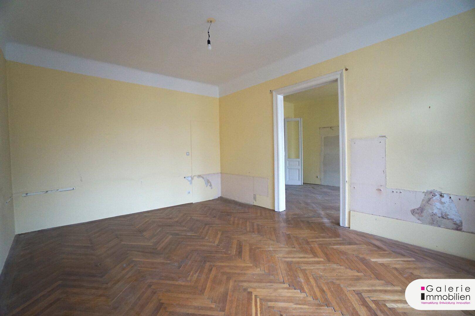 Geräumiges Zimmer mit Fischgrätparkett und Spuren an den Wänden, bereit für Renovierung.