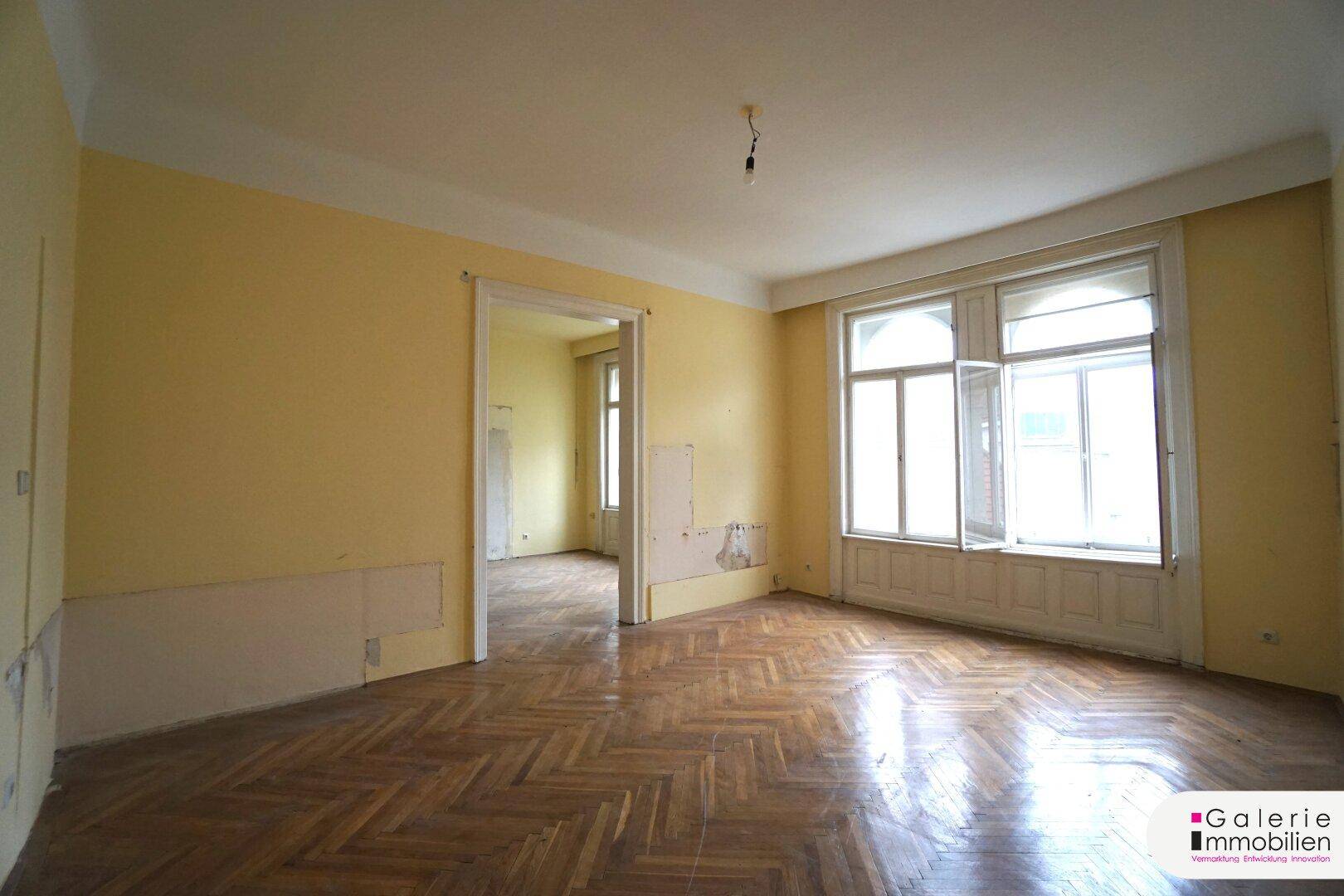 Geräumiges Wohnzimmer mit Fischgrätparkett, hohen Fenstern und Renovierungsbedarf.