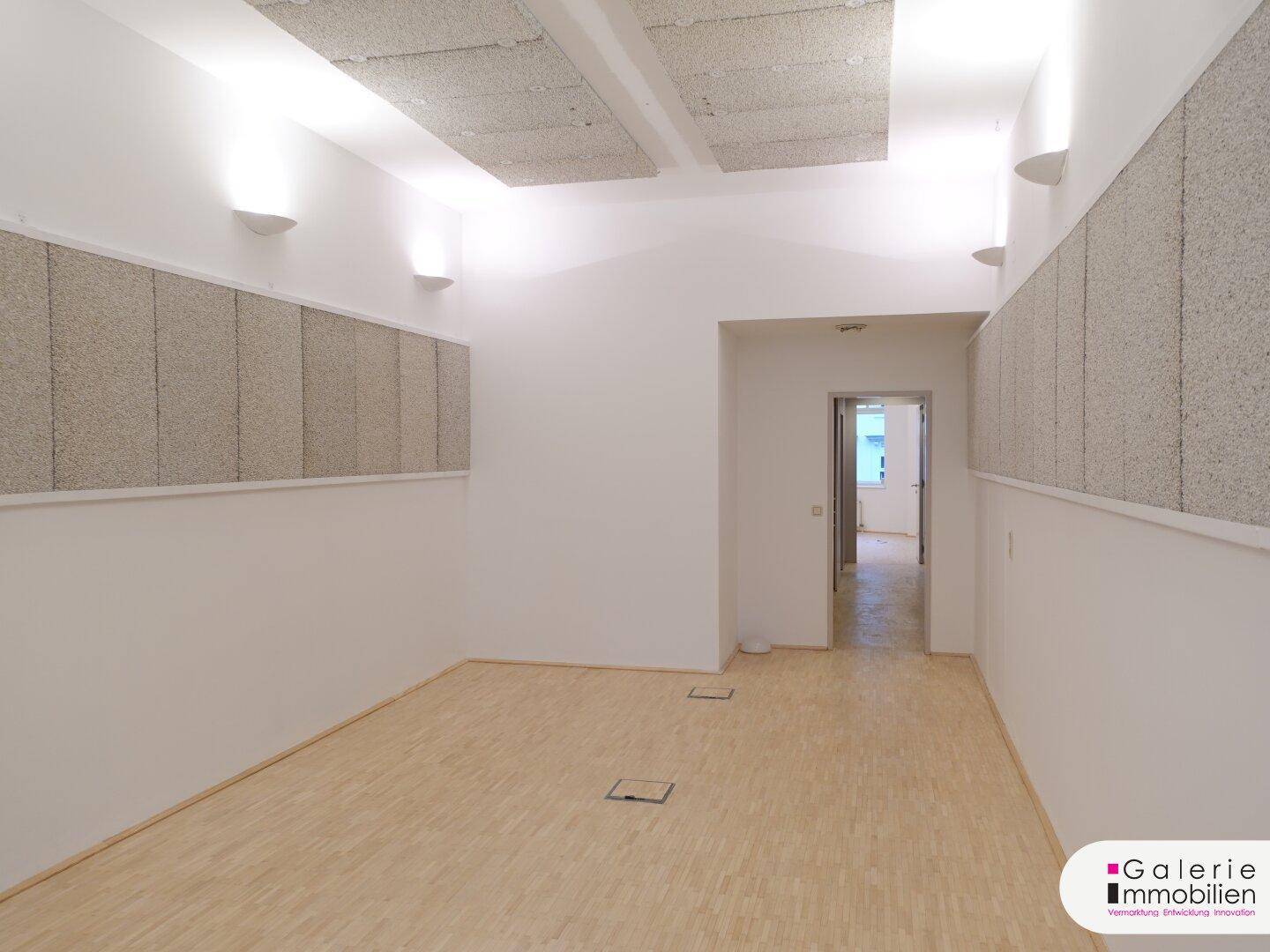 Heller Raum mit Holzboden, schallabsorbierenden Decken- und Wandpaneelen und Zugang zu einem weiteren Raum.