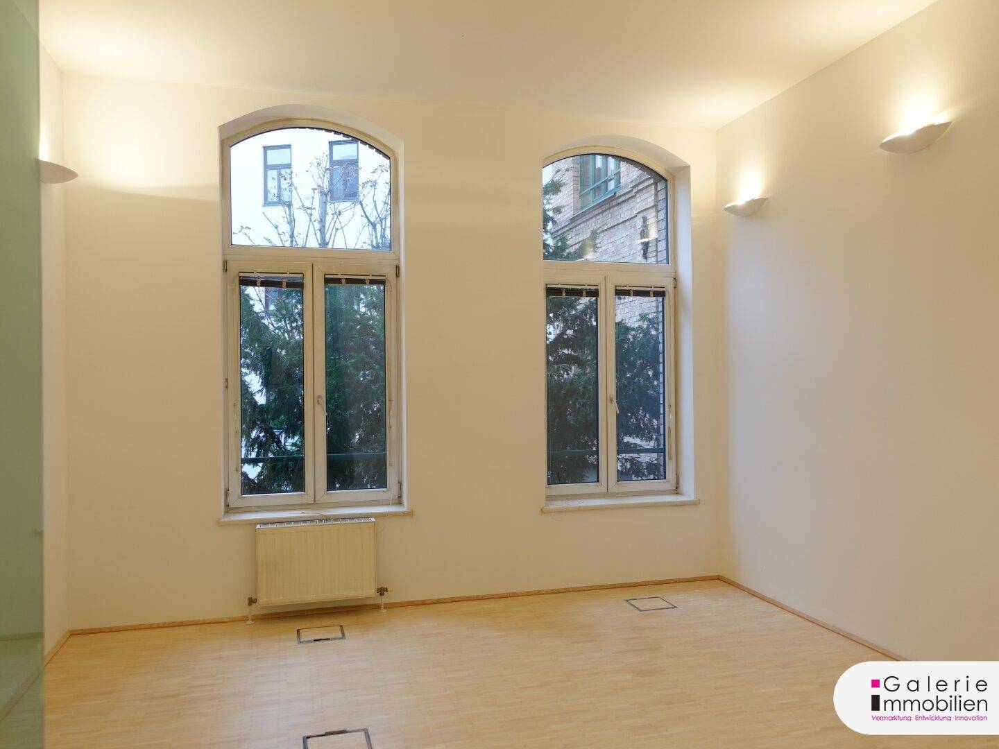 Heller Raum mit zwei hohen Bogenfenstern und Holzboden.