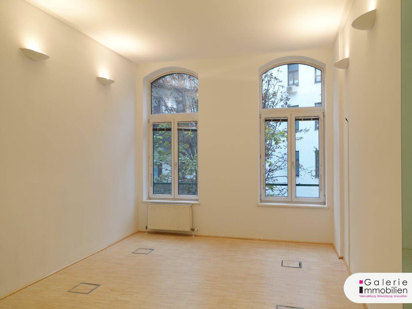 Geräumiger, heller Raum mit zwei hohen Bogenfenstern und Holzboden.