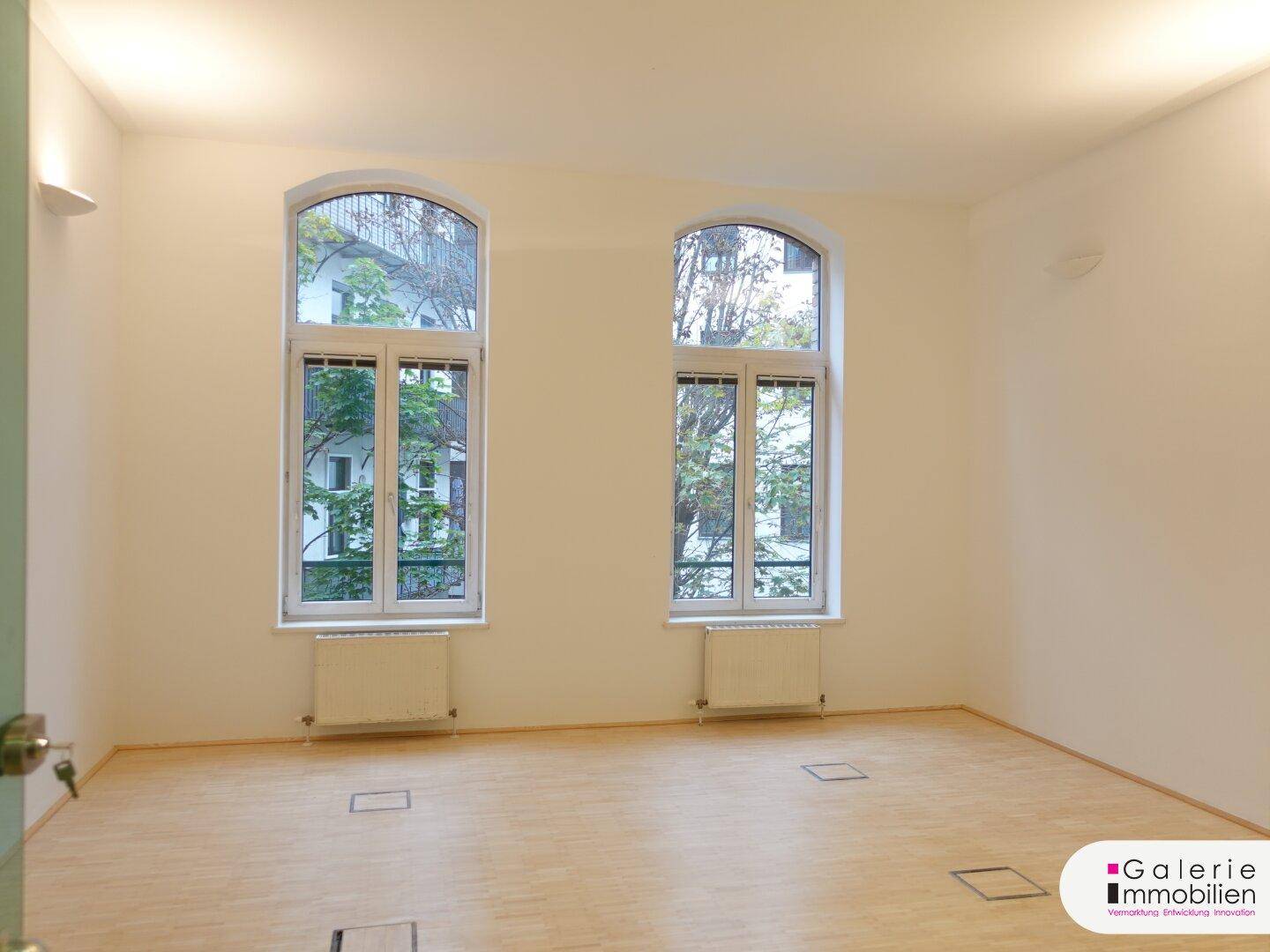 Heller Raum mit zwei hohen Bogenfenstern und Holzboden.