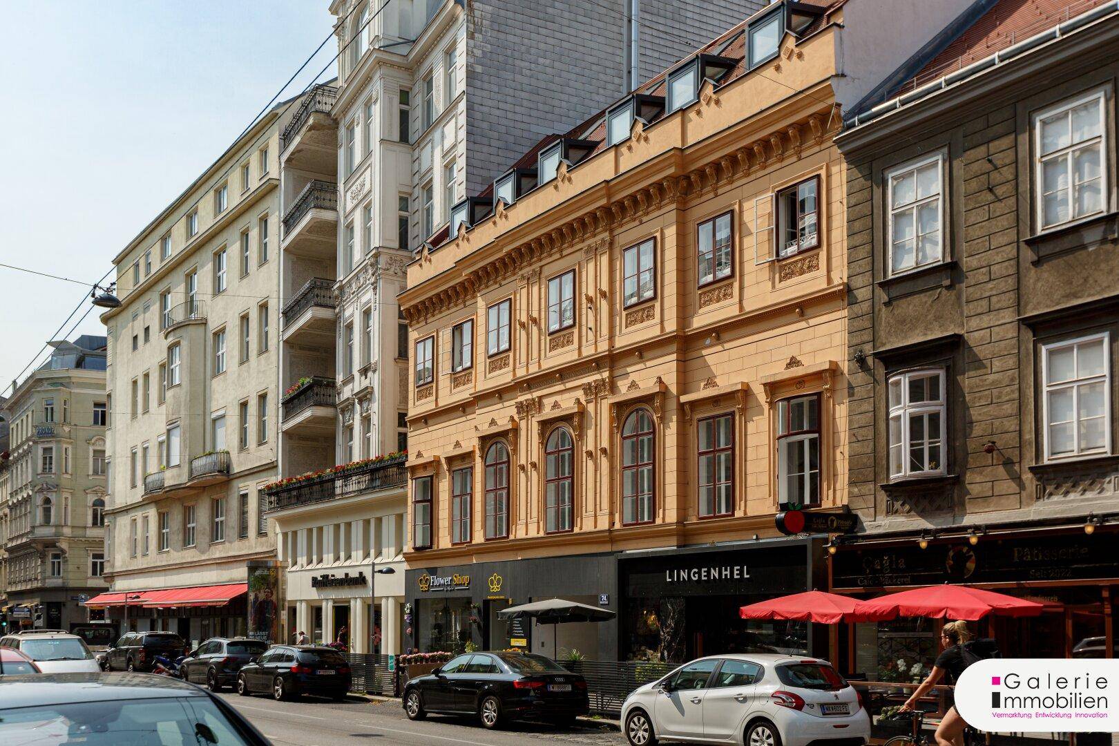 Historische Fassade eines Gebäudes an einer belebten Straße mit Geschäften im Erdgeschoss und Dachgauben.