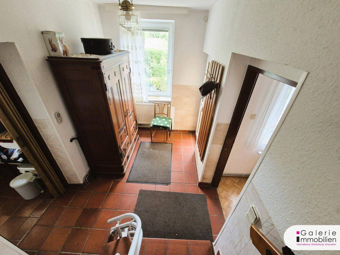 Eingangsbereich mit Terrakotta-Fliesenboden, Holzschrank und Fenster mit Blick ins Grüne.